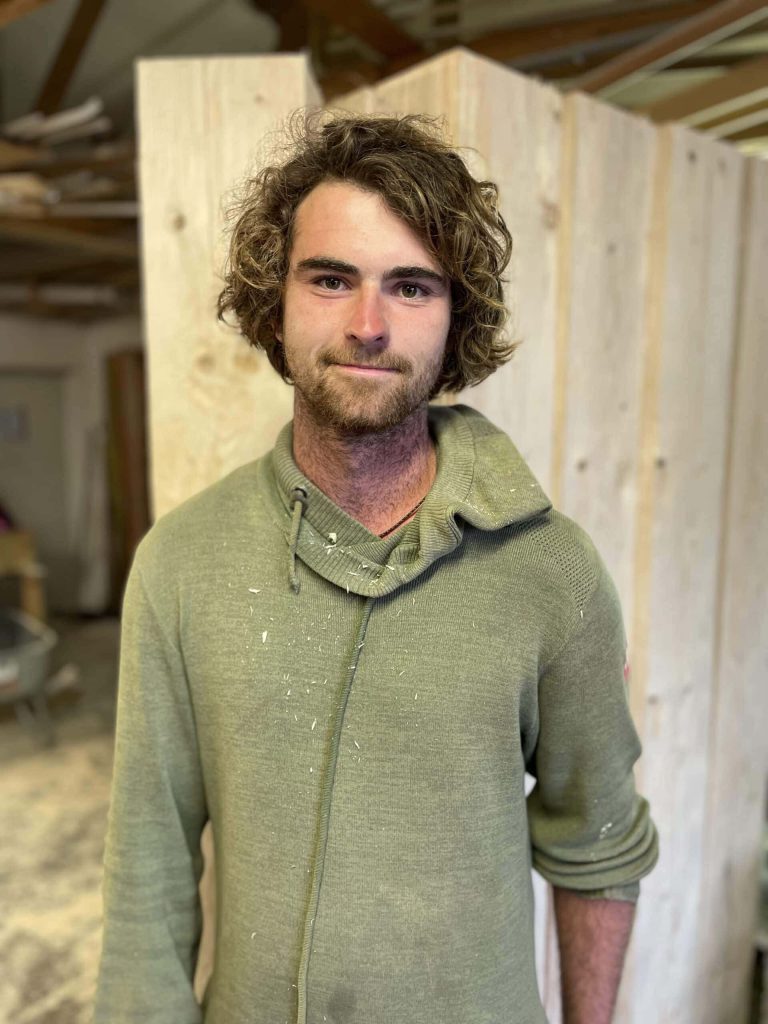 Junger Handwerker in einem Holzbauunternehmen mit Holzstücken im Hintergrund.