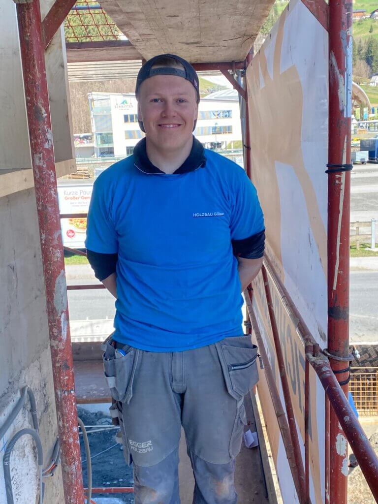 Zimmerer von Holzbau Gläser auf einem Baugerüst in Piesendorf, lächelnd in Arbeitskleidung.