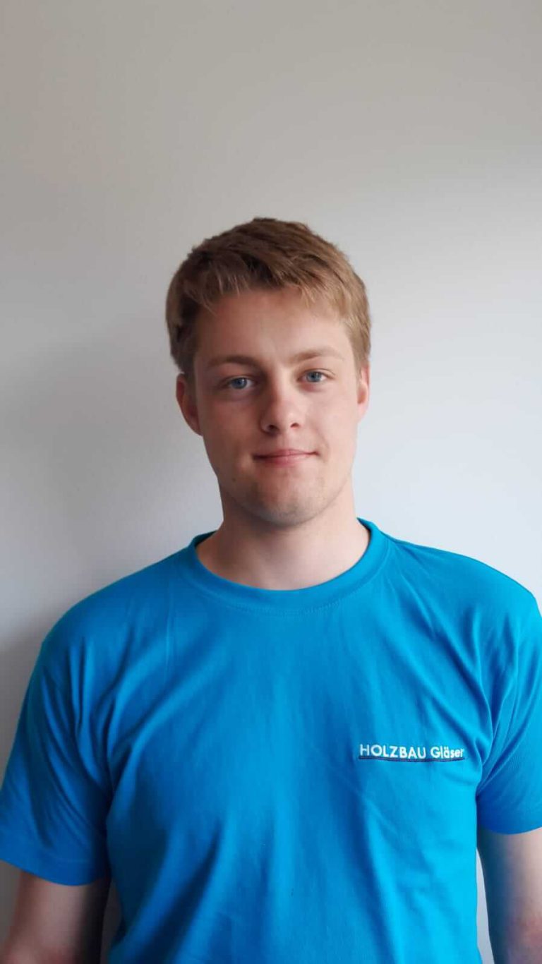 Zimmerer-Lehrling von Holzbau Gläser, lächelnd in blauem T-Shirt vor neutralem Hintergrund.
